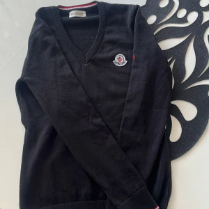 Svart Moncler stickad tröja S - Svart stickad tröja från Moncler med v-ringning och broderad logga på bröstet. Långa ärmar och ribbade muddar med röd och vit detalj vid halsen. Perfekt för dig som gillar stilrena och klassiska plagg med premiumkänsla.