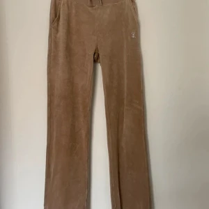 Beiga velourbyxor Juicy Couture S - Supermysiga beiga velourbyxor från Juicy Couture med strassdetaljer och Juicy-logga på baksidan. Byxorna har normal passform, raka ben och resår i midjan. Perfekta för en chill och trendig look. Storlek: S Behöver tvättas 