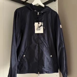 Mörkblå vindjacka från Moncler - Snygg mörkblå vindjacka från Moncler med huva och dragsko, klassisk logga på ärmen och två fickor framtill med tryckknappar. Jackan har hel dragkedja och sportig känsla, perfekt för dig som gillar stilren och clean look.