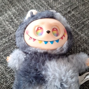 Labubu  - Bilderna visar en mjuk leksak med färgglada ögon och ett brett leende, klädd i en lurvig grå dräkt. Det finns ingen bok synlig på bilderna, endast en tagg med texten 'THE MONSTERS'. Ingen information om boktitel eller författare kan identifieras.