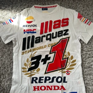 Repsol Honda Marquez MotoGP t-shirt - Vit t-shirt med stora tryck fram och bak från Repsol Honda och Marc Marquez MotoGP 2014 World Champion. Flera loggor i rött, svart, blått och guld, samt detaljerade sponsortryck på ärmarna. Klassisk passform och crewneck. Perfekt för racingfans!