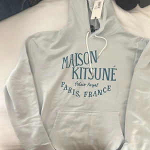 Ljusblå Maison Kitsuné hoodie - Snygg ljusblå hoodie från Maison Kitsuné med tryck i blått framtill. Klassisk känguruficka, vit snörning i huvan och mjuk bomullskänsla. Perfekt för dig som gillar streetwear och fransk stil.