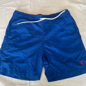 Blå badshorts Polo Ralph Lauren  - Snygga blå badshorts från Polo Ralph Lauren i storlek M med vit snörning i midjanoch  röd broderad logga på benet. Insidan har rött foder och vitt meshfoder för extra komfort. Perfekta för stranden eller poolen!