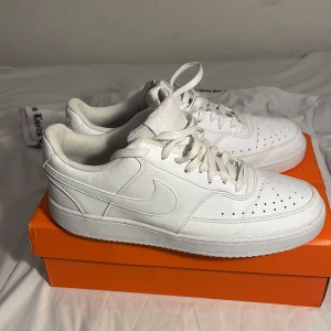 Nike Air Force 1 helvita sneakers - Fräscha helvita Nike Air Force 1 sneakers i klassisk low-modell. Skorna har snyggt skinnmaterial, perforerade detaljer på tån och ikonisk swoosh på sidorna. Perfekta för dig som gillar clean och stilren streetwear.