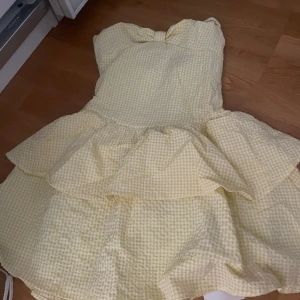 Gul rutig kortklänning från Cider - Supersöt gul och vit rutig kortklänning från Cider med volanger och axelbandslös design. Klänningen har en stor rosett framtill och smockad rygg för extra komfort. Perfekt för sommarens alla tillfällen!