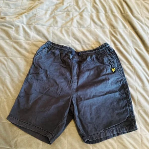 Mörkblå shorts från Lyle & Scott Junior - Snygga mörkblå shorts från Lyle & Scott Junior med resår och snörning i midjan. Shortsen har sidofickor, bakfickor och den klassiska gula loggan broderad på vänster sida. Perfekta för sommardagar!
