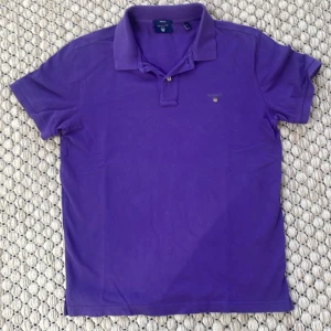 Lila piké t-shirt från GANT - Säljer en lila piké t-shirt från GANT i klassisk modell med krage och knappar. T-shirten har korta ärmar och broderad GANT-logga på bröstet. Perfekt för en stilren och avslappnad look. Storlek:M. 