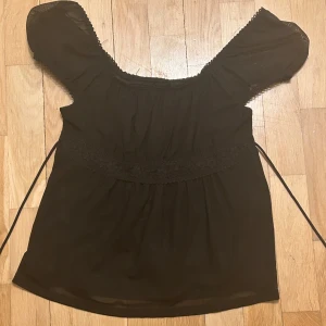 Svart blus med spetsdetaljer - Säljer en svart blus med vackra spetsdetaljer runt midjan och vid halsringningen. Blusen har korta puffärmar och knytband i midjan för en fin passform. Perfekt för dig som gillar romantisk stil.