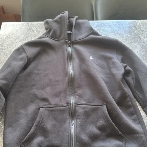 Svart hoodie med dragkedja från Polo Ralph Lauren - Svart hoodie från Polo Ralph Lauren med dragkedja framtill och klassisk broderad logga på bröstet. Tröjan har huva och två fickor framtill. Perfekt för en avslappnad och stilren look.