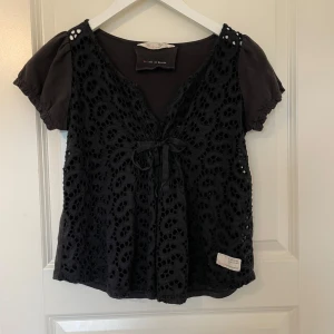 Svart blus från Odd Molly - Supersöt svart blus från Odd Molly med korta ärmar och v-ringning. Framsidan har ett vackert spetsmönster och en knytning vid bysten. 