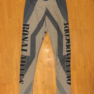Blåvita träningstights med textmönster M - Säljer ett par blåvita leggings med grafiskt mönster och stora tryckta texter som 'ONLYHARDCORE' och 'RONALADIES' längs benen. Tillverkade i stretchigt material som består av polyester och spandex. Perfekta för dig som vill sticka ut på gymmet eller i vardagen. 95% polyester 5% spandex  Beninnerlängd: 75 m Midja: 72 cm Resår i midja