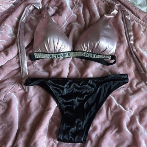 Rosa och svart bikini från Victoria's Secret - Säljer en snygg bikini från Victoria's Secret med glittrig rosa överdel och svarta trosor. Överdelen har knyt i nacken och glittrig detalj med text under bysten. Materialet är glansigt och känns lyxigt.
