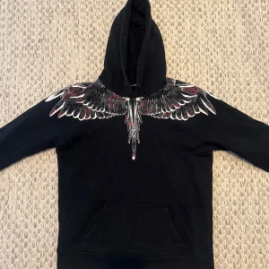 Marcelo Burlon hoodie - Säljer en svart hoodie från Marcelo Burlon med ikoniskt vingmönster i vitt, rött och grått över axlar och huva. Tröjan har lång ärm, huva och känguruficka framtill. Perfekt för dig som vill sticka ut med en unik streetwear-stil.