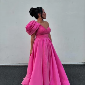 Rosa långklänning - Elegant rosa långklänning med en voluminös oneshoulder-ärm. Klänningen har en vacker drapering över bysten och en vid kjol som ger en fantastisk silhuett. Perfekt för balen eller bröllop
