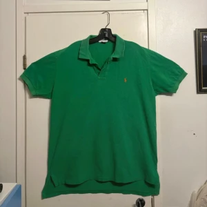 Grön pikétröja från Polo Ralph Lauren - Säljer en klassisk grön pikétröja från Polo Ralph Lauren med orange broderad logga på bröstet. Tröjan har korta ärmar, krage och knappar vid halsen. Tillverkad i 100% bomull för en bekväm känsla.