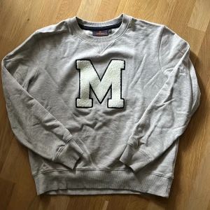 Grå sweatshirt från MORRIS - Säljer en grå sweatshirt med ett stort, fluffigt M på bröstet. Tröjan har rund halsringning och långa ärmar med ribbade muddar. Perfekt för en avslappnad stil. Unisex. Använd 1 ggr. Fin melange färg. 