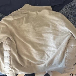 Moncler Cardigan - Säljer en beige jacka från moncler med quiltade partier framtill och på sidorna. Jackan har hög krage, dragkedja och är långärmad. Perfekt för kyligare dagar och har en sportig look.