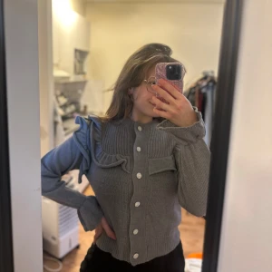 Grå kofta med volangdetaljer och knappar - Säljer en grå kofta med volangdetaljer vid axlar och bröst, samt vita knappar framtill. Koftan har lång ärm och ribbade muddar. Perfekt för dig som vill ha något unikt och stilrent i garderoben.