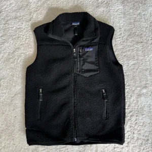 Svart fleeceväst från Patagonia - Svart fleeceväst från Patagonia med dragkedja framtill och tre fickor med dragkedja, varav en på bröstet. Västen har hög krage och är tillverkad i mjuk fleece med detaljer i syntetmaterial.