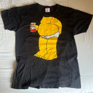 Simpson T-shirt - Cool svart t-shirt från Sama med en stor gul tecknad figur som håller en ölburk på framsidan. T-shirten har rund hals och korta ärmar. Perfekt för dig som gillar unika och roliga tryck!