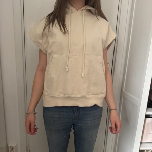 Beige ärmlös hoodie - Säljer en beige hoodie med korta ärmar och huva. Tröjan har en magficka och dragsko vid huvan. Perfekt för en avslappnad stil och passar bra till jeans eller leggings.