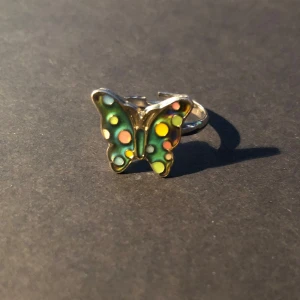 Färgändrande Fjärilsring i silverfärg med färgglada prickar - Söt ring med en fjäril i silverfärg. Den ändrar färg beroende på temperaturen på ringen. Fjärilen har prickar i grönt, gult, rosa och blått. Justerbar storlek så den passar de flesta. Perfekt accessoar för dig som gillar färg och lekfulla detaljer.