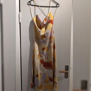 Färgglad klänning från Stradivarius - Säljer en långklänning från Stradivarius med smala korsade axelband och v-ringad draperad urringning. Klänningen har ett abstrakt mönster i gult, orange, vitt och lila på glansigt satinliknande material. 