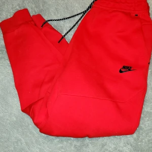 Röda mjukisbyxor från Nike - Snygga röda mjukisbyxor från Nike med svart logga på benet och dragsko i midjan. Byxorna har en avslappnad passform och är perfekta för en sportig stil. Tillverkade i mjukt material för extra komfort.