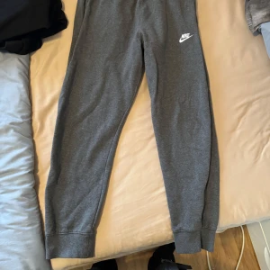 Grå mjukisbyxor från Nike med hoodie - Gråa mjukisbyxor från Nike med resår i midjan och muddar vid bensluten. Byxorna har en liten vit Nike-logga på vänster lår och en bakficka. Perfekta för chill eller träning. Med hoodie 