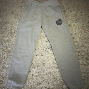 Grå mjukisbyxor från Canada Goose - Säljer ett par grå mjukisbyxor från Canada Goose med broderad logga på ena benet. Byxorna har resår i midjan med snörning och muddar vid bensluten. Perfekta för en avslappnad stil.