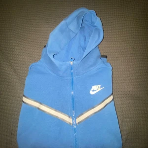 Nike tech  - Snygg blå hoodie från Nike med dragkedja framtill och huva. Tröjan har vita och beige ränder över bröstet och en liten Nike-logga på vänster sida. Perfekt för en sportig och avslappnad stil.