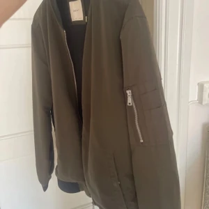 Olivgrön bomberjacka från Celio* - Säljer en olivgrön bomberjacka från Celio* med klassisk passform. Jackan har dragkedja framtill, ribbade muddar och krage i svart samt en ärmficka med dragkedja. Insidan är fodrad med mesh för extra komfort.