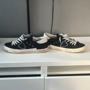 Svarta Philippe Model sneakers med vita detaljer - Säljer ett par svarta Philippe Model sneakers med vita snören och vit häl. Skorna har en klassisk låg modell med rund tå och platt sula. Märkeslogga på sidan och bak på hälen. Perfekta för en stilren vardagslook.