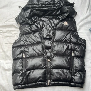 Svart Moncler dunväst med luva  - Säljer en svart dunväst från Moncler med avtagbar huva och två dragkedjefickor framtill. Den är ej äkta. Säljer pga att jag inte använder den. Priset är inte hugget i sten!