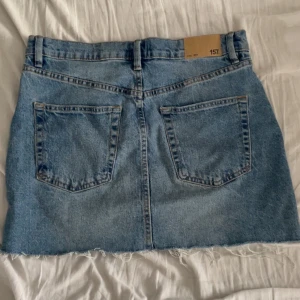 Blå jeanskjol med rå kant - Säljer en klassisk blå jeanskjol med hög midja och rå, fransig nederkant. Kjolen har två bakfickor, framfickor och knappar framtill. Perfekt för en avslappnad stil.