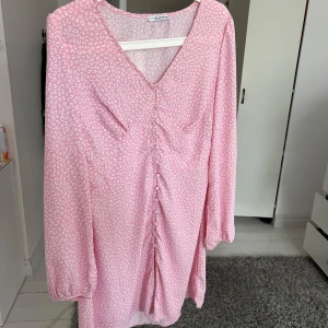 Rosa mönstrad klänning med v-ringning från NA-KD - Säljer en söt rosa klänning från Nelly med vitt mönster, v-ringning och långa puffärmar. Klänningen har knappar och rynkdetalj framtill som ger en fin form. Perfekt för vår och sommar! Den är i storlek 40 men lite insydd i midjan! Skulle säga att den är som en 38🌸💗