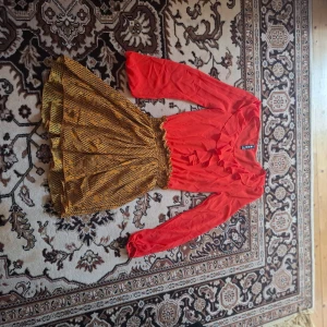 Röd och gul klänning med volangdetaljer - Säljer en färgstark klänning med röd långärmad överdel och gulmönstrad kjol. Klänningen har volangdetaljer framtill och resår i midjan för en bekväm passform. Perfekt för dig som vill sticka ut!