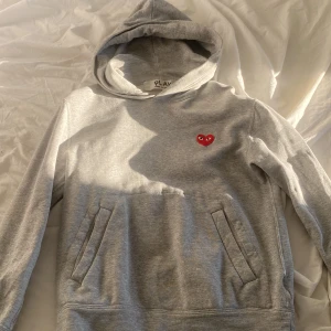 Grå hoodie från Comme des Garçons Play - Grå hoodie från Comme des Garçons Play med den ikoniska röda hjärt-loggan broderad på bröstet. Tröjan har huva, magficka och långa ärmar. Perfekt för en avslappnad och stilren look.
