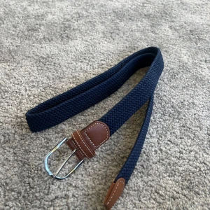 Mörkblått flätat bälte med bruna skinn-detaljer - Snyggt mörkblått flätat bälte med detaljer i brunt skinn och silvrigt spänne. Perfekt accessoar för att lyfta din outfit. Bältet har en klassisk design med kontrastsömmar på skinnet.