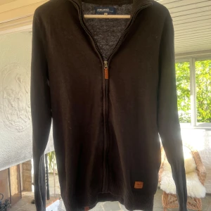 Mörkgrå ziptröja från Blend - Säljer en mörkgrå långärmad ziptröja från Blend i storlek M. Tröjan har hel dragkedja framtill, ribbade muddar och en liten läderdetalj vid nederkanten. 
