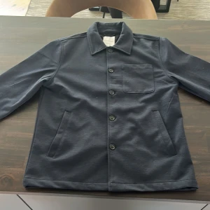Mörkblå overshirt från Lindbergh - Säljer en stilren mörkblå overshirt från Lindbergh i regular fit. Jackan har klassisk krage, tre fickor framtill och knäppning med knappar. Perfekt att slänga över en t-shirt för en clean look. Pris kan diskuteras.