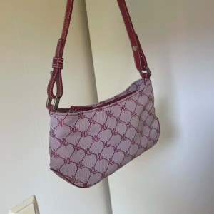 Röd och vit axelväska från Lauren Ralph Lauren - Säljer en snygg axelväska från Lauren Ralph Lauren i rött och vitt med mönster och matchande röd axelrem. Väskan har dragkedja upptill och logotypmönster över hela ytan. Perfekt accessoar för att lyfta din outfit!
