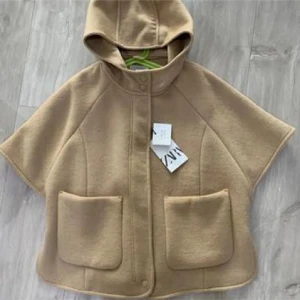 Beige capejacka med huva från ZARA - Säljer en beige capejacka från ZARA med huva och stora framfickor. Jackan har knäppning framtill och är i ett mjukt material som känns lyxigt. Perfekt för lager-på-lager och en snygg detalj till din outfit. I barn storlek men passar xs/s
