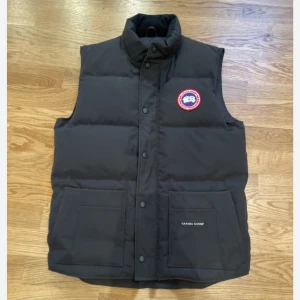 Svart dunväst från Canada Goose, påse följer med  - Säljer en svart dunväst från Canada Goose med hög krage och tryckknappar framtill. Västen har två stora fickor framtill och en rund logotyp-patch på bröstet. Perfekt för kyliga dagar.köpt i Spanien , påse medföljer ! Är äkta !