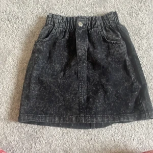 Svart jeanskjol med resår från H&M - Säljer en svart jeanskjol från H&M med resår i midjan och två fickor fram. Kjolen har en lätt tvättad look och knapp framtill. Perfekt att matcha med en enkel topp eller hoodie.