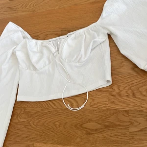 Vit croppad blus med puffärmar - Säljer en vit croppad blus med långa puffärmar och knytning framtill. Toppen har en söt, feminin stil och är tillverkad i ett lätt, strukturerat tyg. Perfekt att styla med jeans eller kjol.