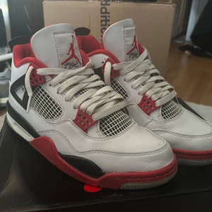 Nike Air Jordan 4 Fire Red - Säljer ett par Nike Air Jordan 4 Fire Red med klassisk vit läderöverdel, svarta och röda detaljer samt meshpaneler på sidorna. Skorna har tydlig Nike Air-logga på hälen och röd Jumpman på plösen. Perfekta för dig som gillar retrostil och streetwear. Köpta här på Plick och rengörs naturligtvis innan försäljning!