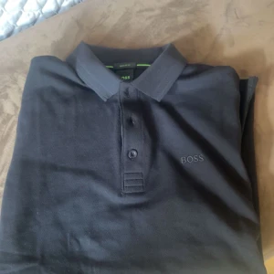 Svart piké t-shirt från boss - Snygg svart piké t-shirt från BOSS med klassisk krage och knappar framtill. Diskret BOSS-logga broderad på bröstet. Perfekt för en stilren och enkel look. Nypris 1100. Helt ny!