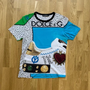 Färgglad t-shirt från Dolce & Gabbana size M  - Unik och färgstark t-shirt från Dolce & Gabbana med grafiska mönster, tryck och broderade detaljer. T-shirten har korta ärmar, svart krage och inslag av blått, grönt, vitt och svart samt dekorativa motiv och text.