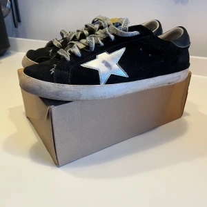Svarta sneakers från Golden Goose med vit stjärna - Snygga svarta sneakers från Golden Goose med vit stjärndetalj på sidan och grå snören med logga. Skorna har platt sula och är tillverkade i mocka med slitna detaljer för en cool look. Skosnören orginal med logga
